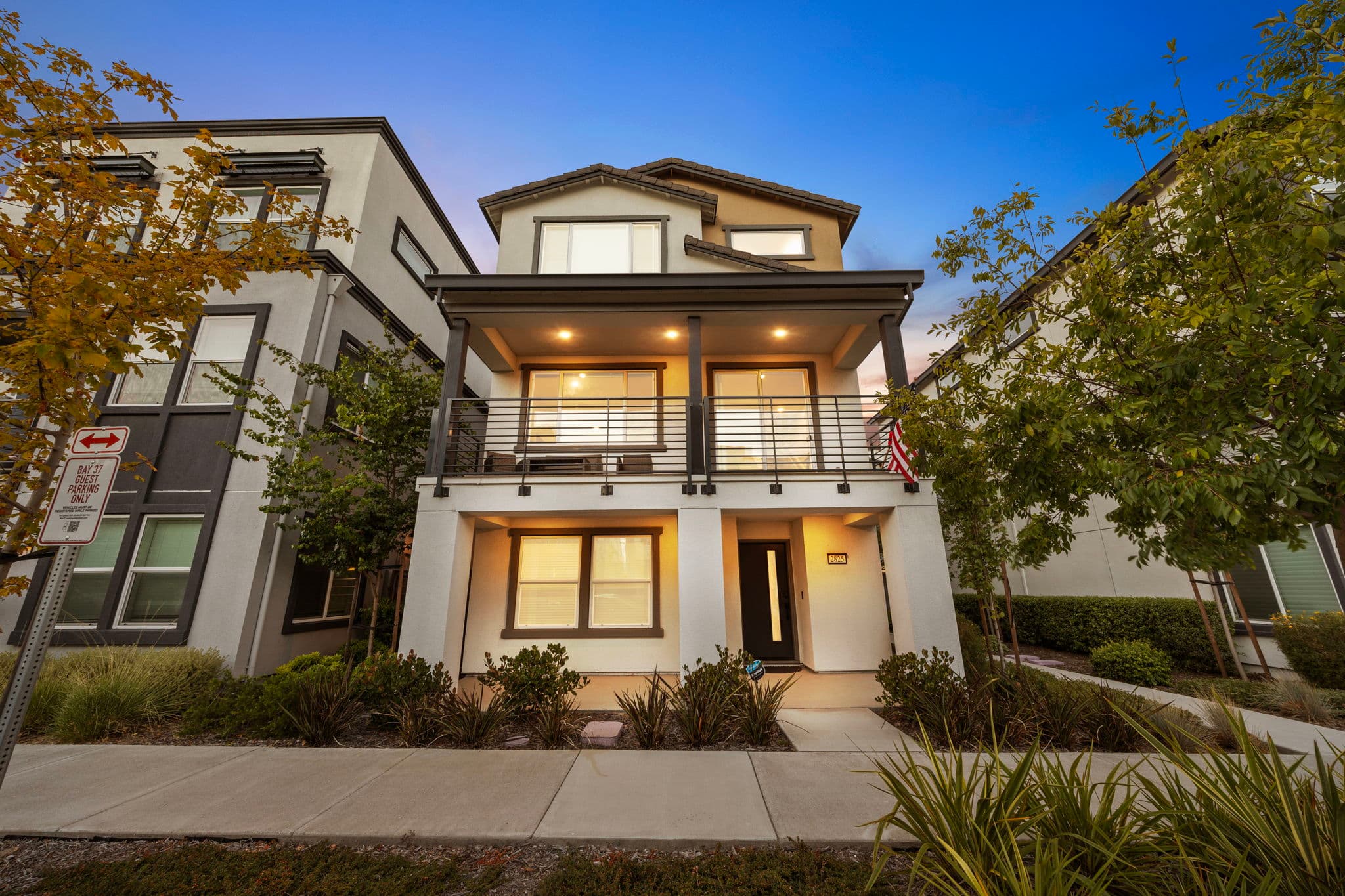 Twilight front elevation — covered porch, full-width balcony, and warm light in every window